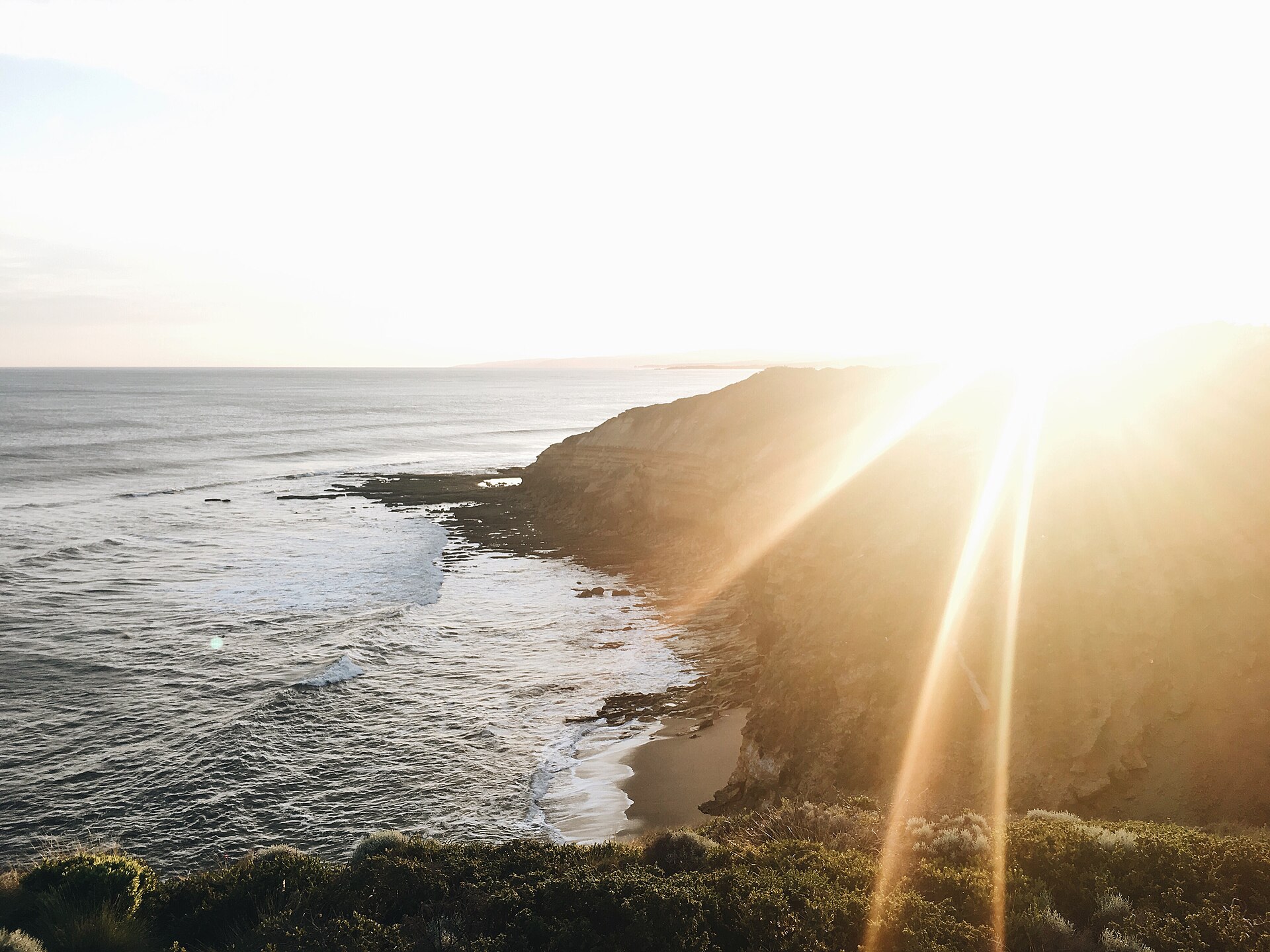Late sun flaring over the clifftop at Bells Beach, with waves breaking along the rocky reef and a small sandy cove below