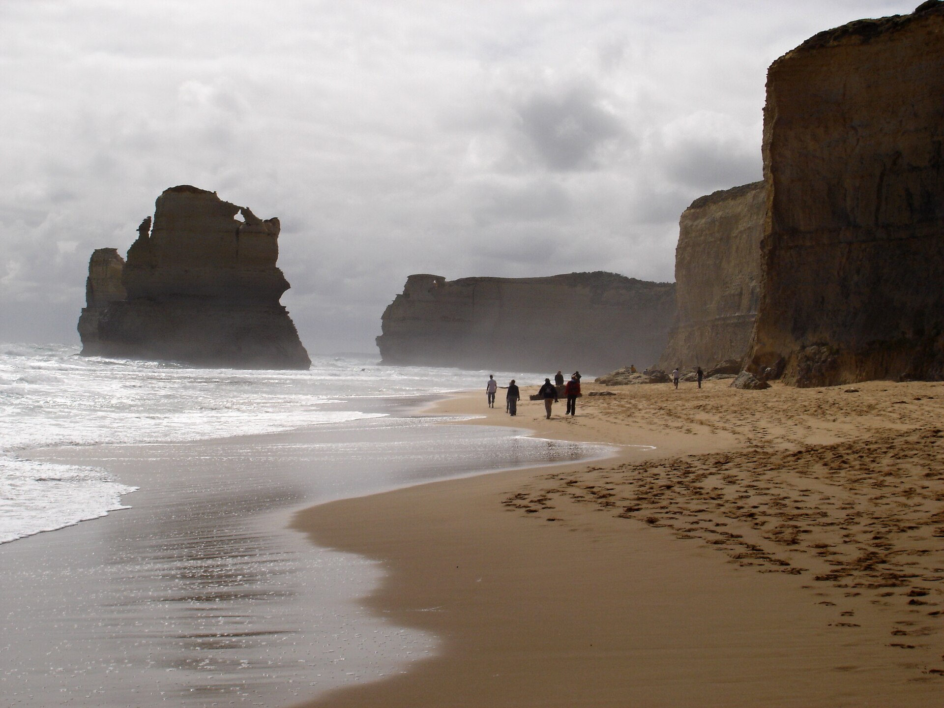 Visitors walking along Gibson Beach beneath tall limestone cliffs, with a large sea stack rising offshore in misty light