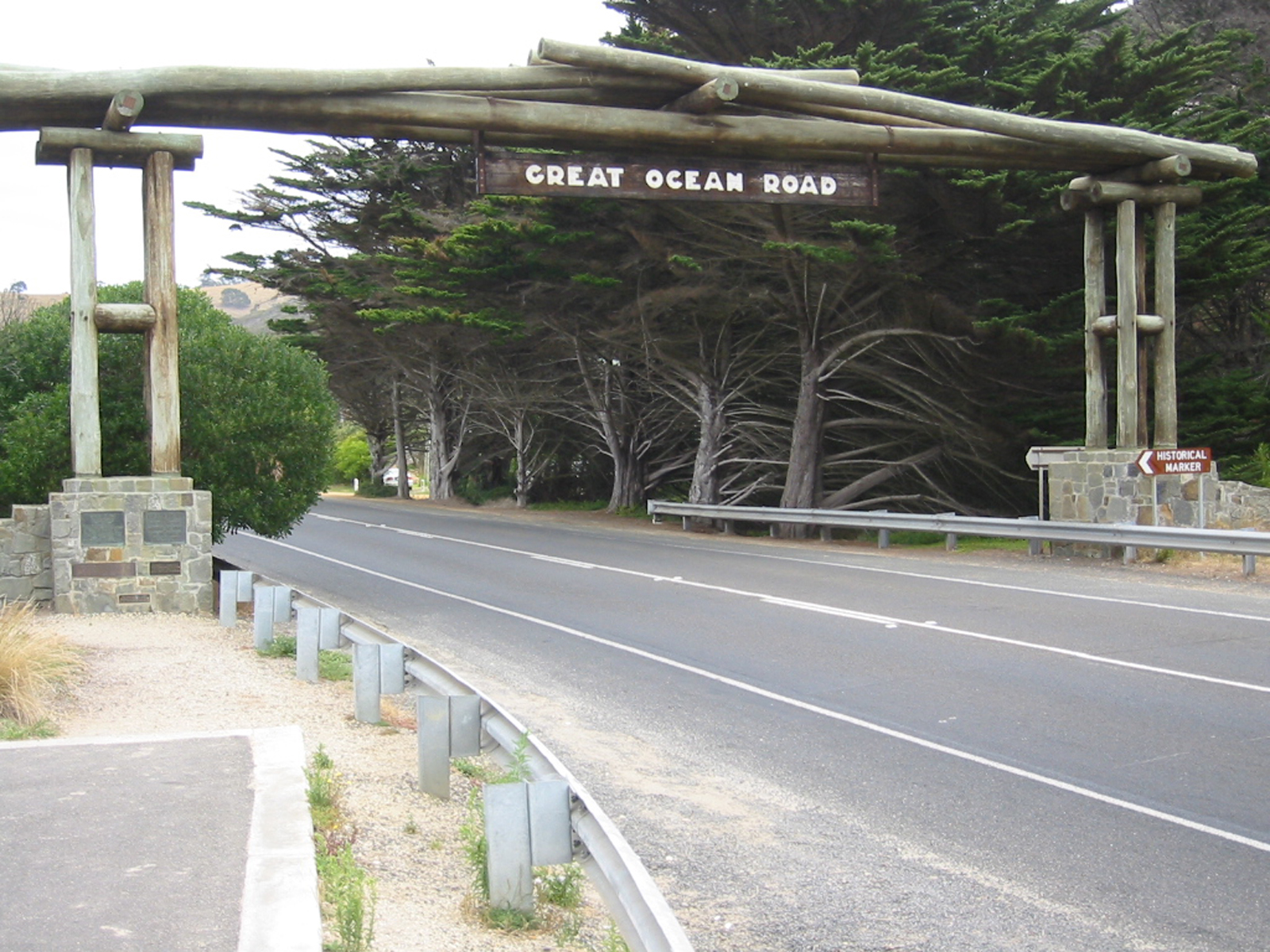 The timber Great Ocean Road memorial arch spanning the road at Eastern View, with stone pillars and cypress trees behind