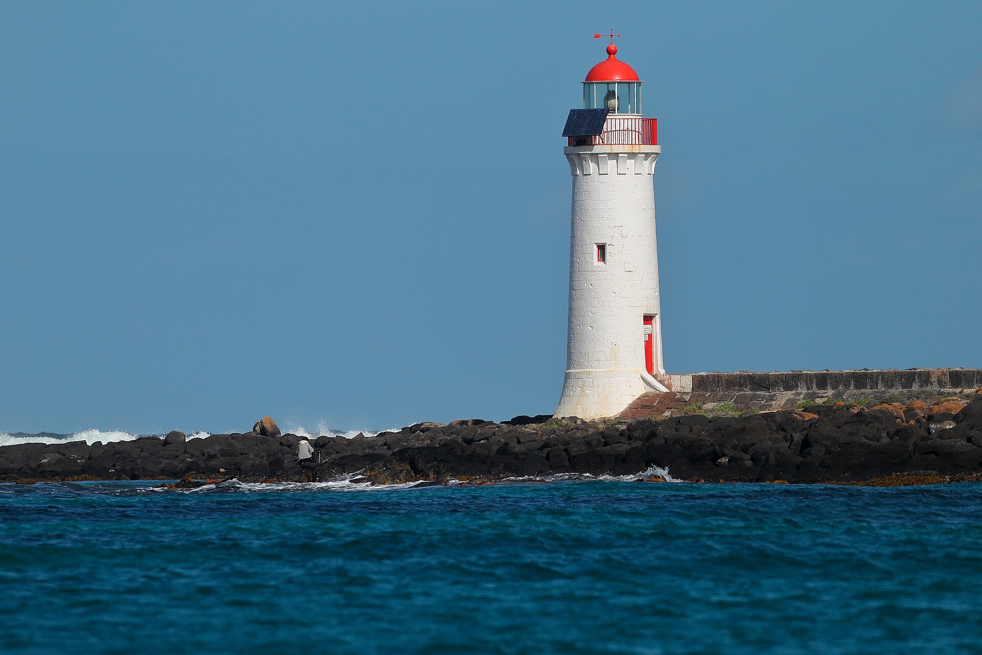 The white Griffiths Island Lighthouse with its red dome on black basalt rocks at Port Fairy