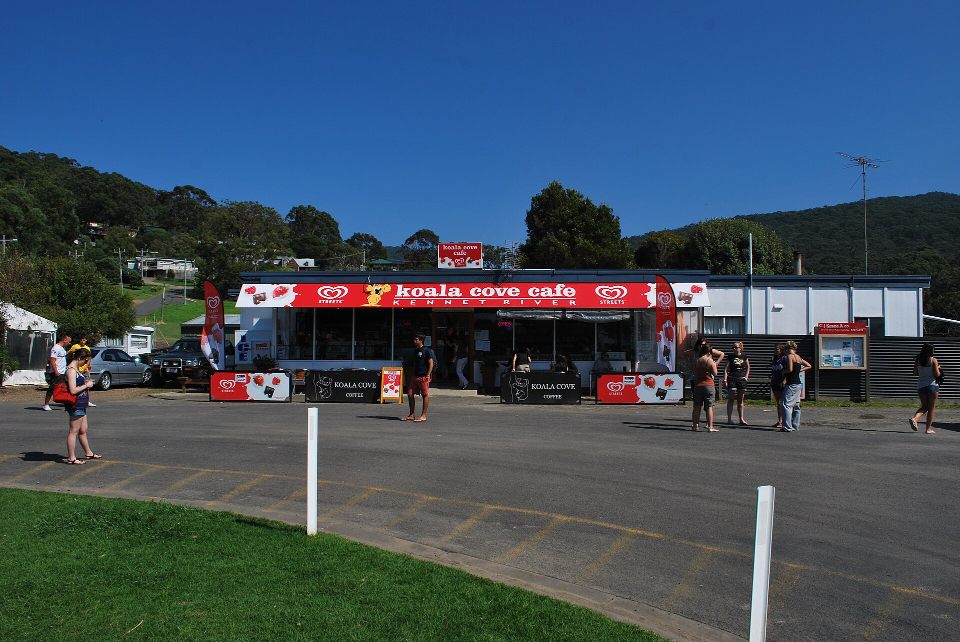 The red-signed Koala Cove Cafe at Kennett River, with people standing in the car park and forested hills behind