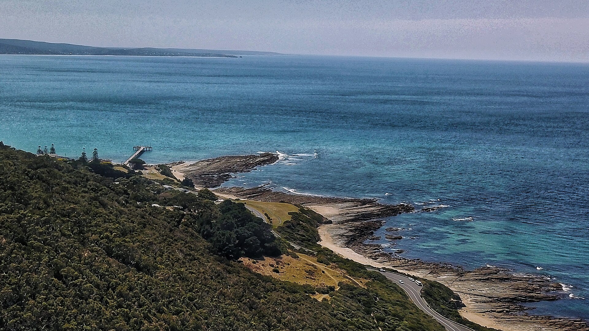 Lorne Pier stretching into turquoise water from a forested headland, with rock platforms and the Great Ocean Road curving along the shore below