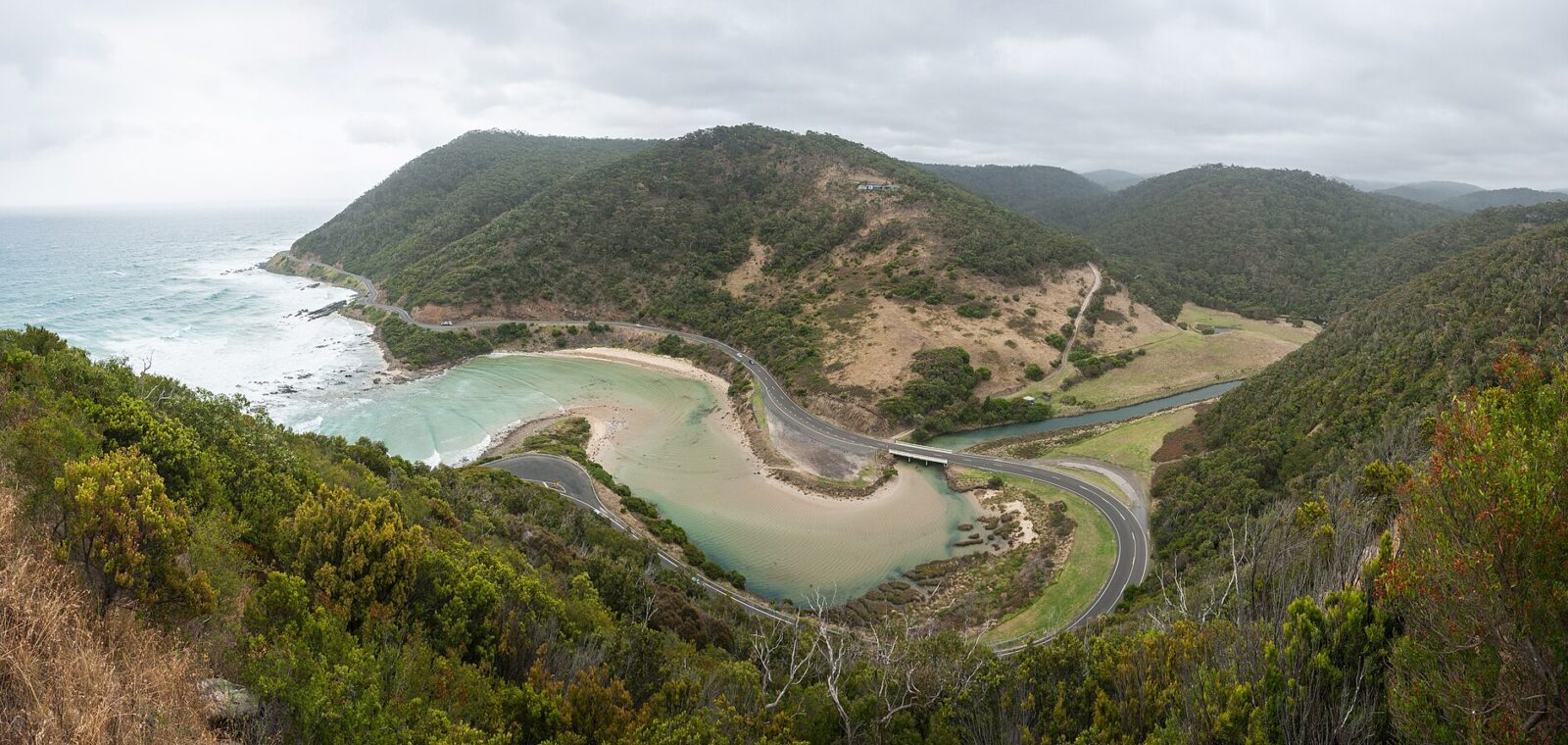 The Great Ocean Road curving around the mouth of the St George River below forested hills, seen from Teddy's Lookout at Lorne
