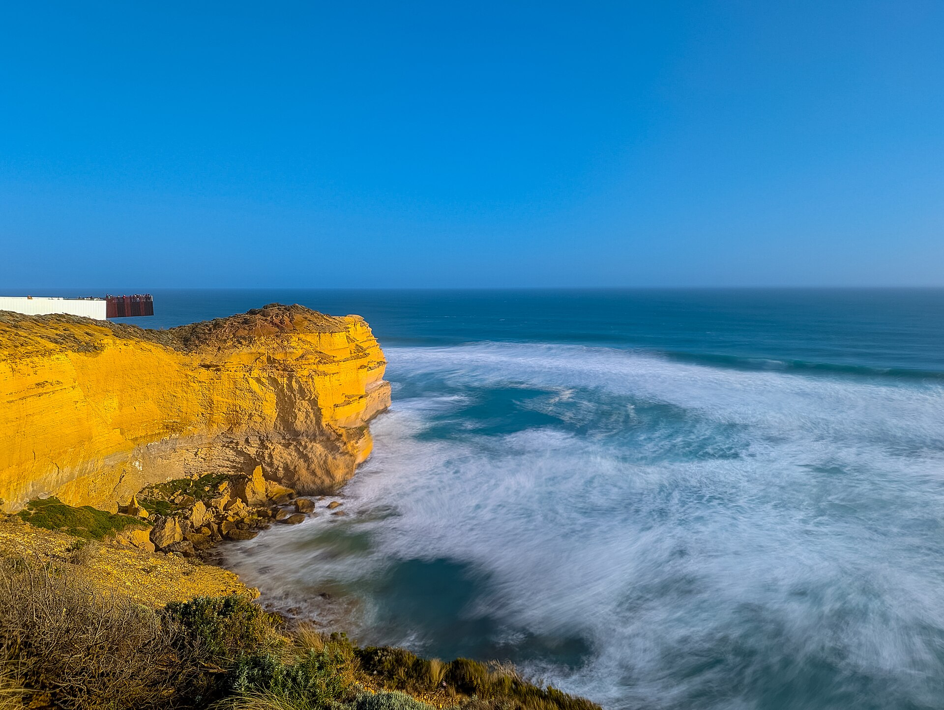 A sheer limestone cliff lit gold by late sun, with long-exposure surf swirling below and a cantilevered viewing platform on the headland