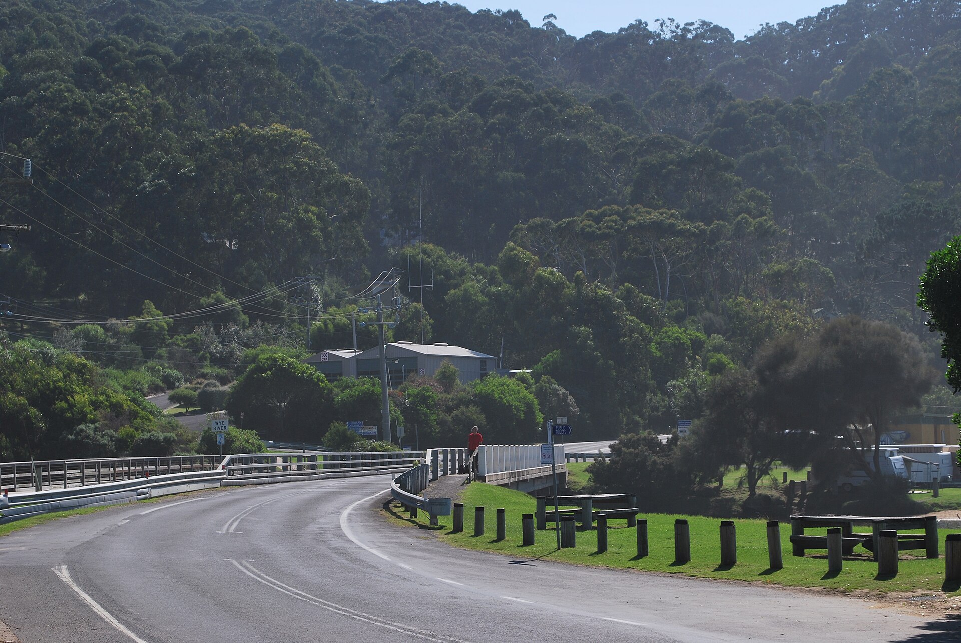 The Great Ocean Road curving over the bridge at Wye River, with forested hills behind and caravans in the holiday park beside the river