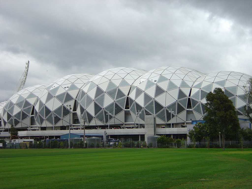 The white geodesic domes of the AAMI Park stadium roof under grey cloud, with a lawn and trees in front
