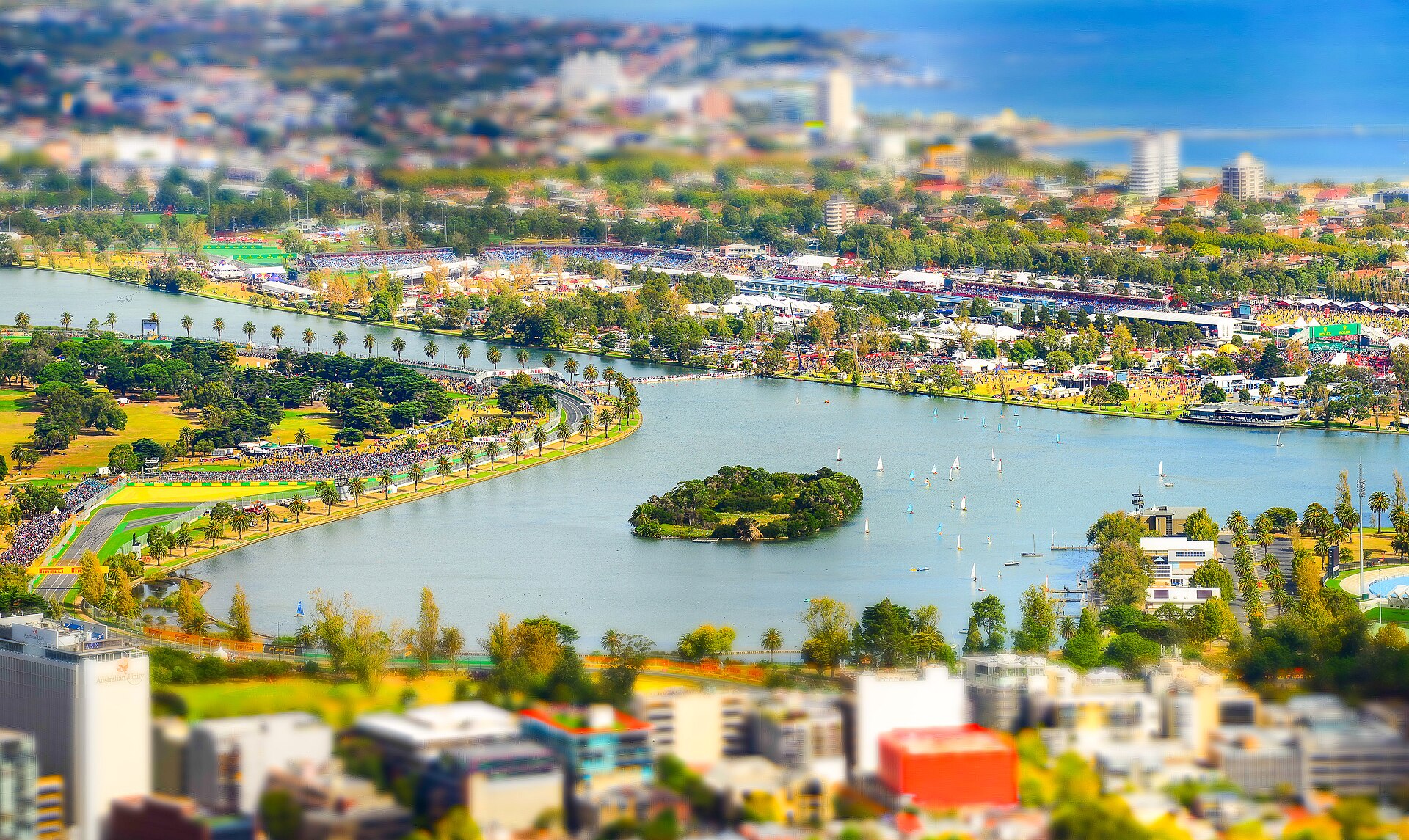 Albert Park Lake seen from high above Southbank, with the Grand Prix circuit, grandstands and parkland around the water and the bay beyond