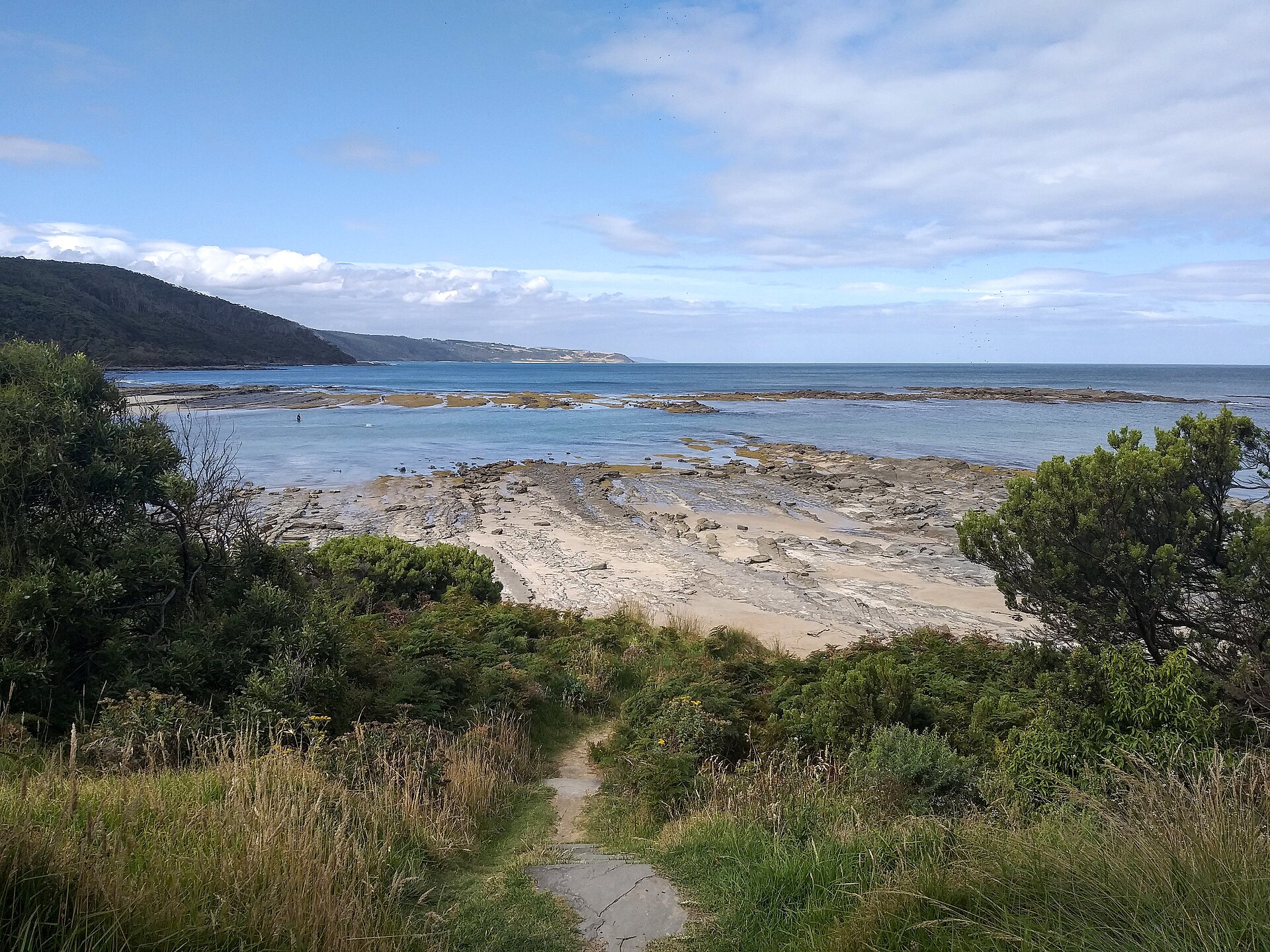 A sandy track through coastal scrub leading down to the flat rock platforms and calm water of Blanket Bay, with forested hills along the coast beyond