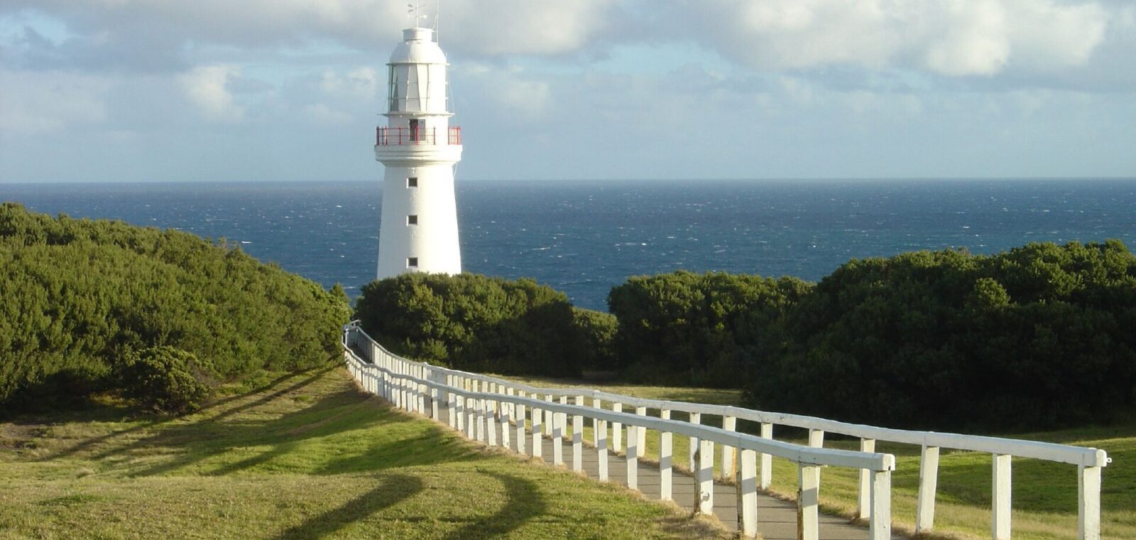 A white post-and-rail fence lines the path across a green lawn to the white Cape Otway lighthouse, with coastal scrub and the ocean behind