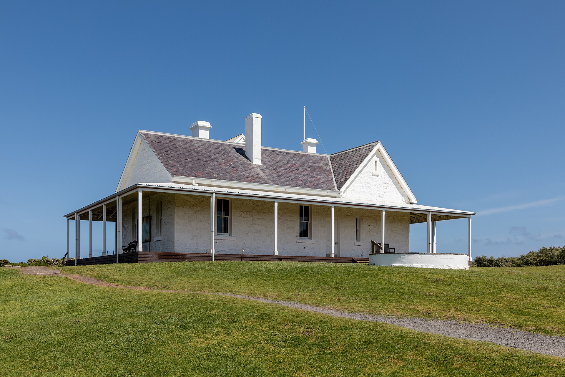 The single-storey Cape Otway Telegraph Station, a white building with a wide verandah and a slate roof, on a lawn under a clear blue sky