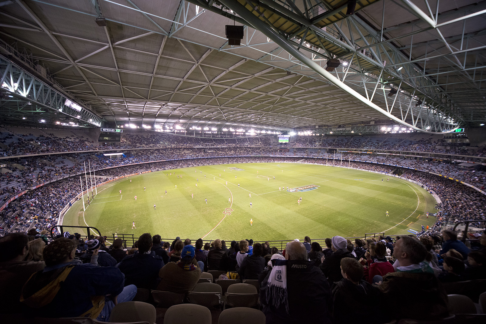 An AFL match under the closed roof of the Docklands stadium, seen from a crowded upper tier with the floodlit oval below