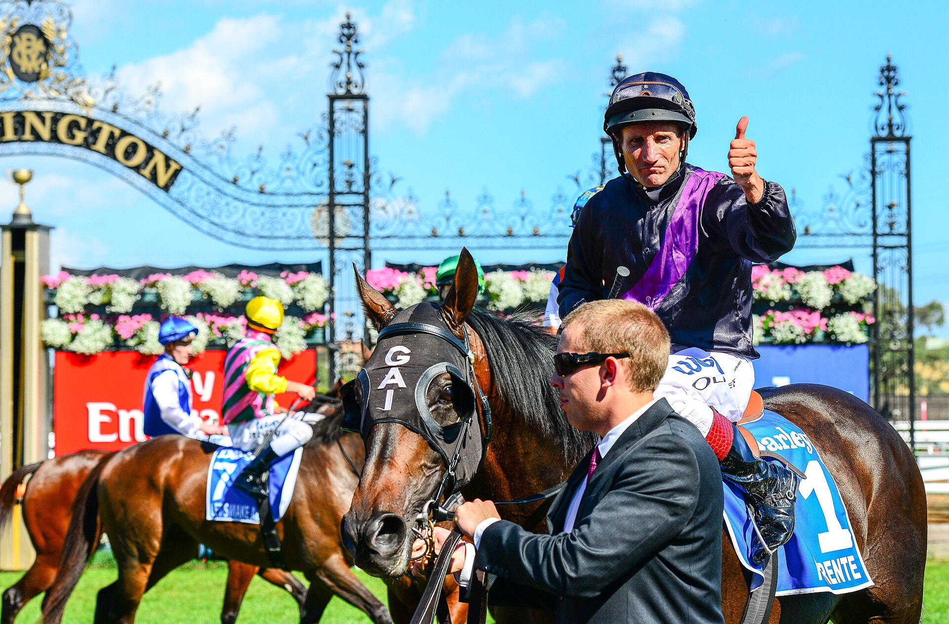 A smiling jockey gives a thumbs-up from a racehorse led by a strapper in sunglasses, with the ornate Flemington gates and a bank of flowers behind