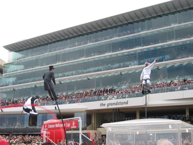 Racegoers on the balconies of The Grandstand at Flemington, with stilt performers in top hats and a red Melbourne Cup balloon in front