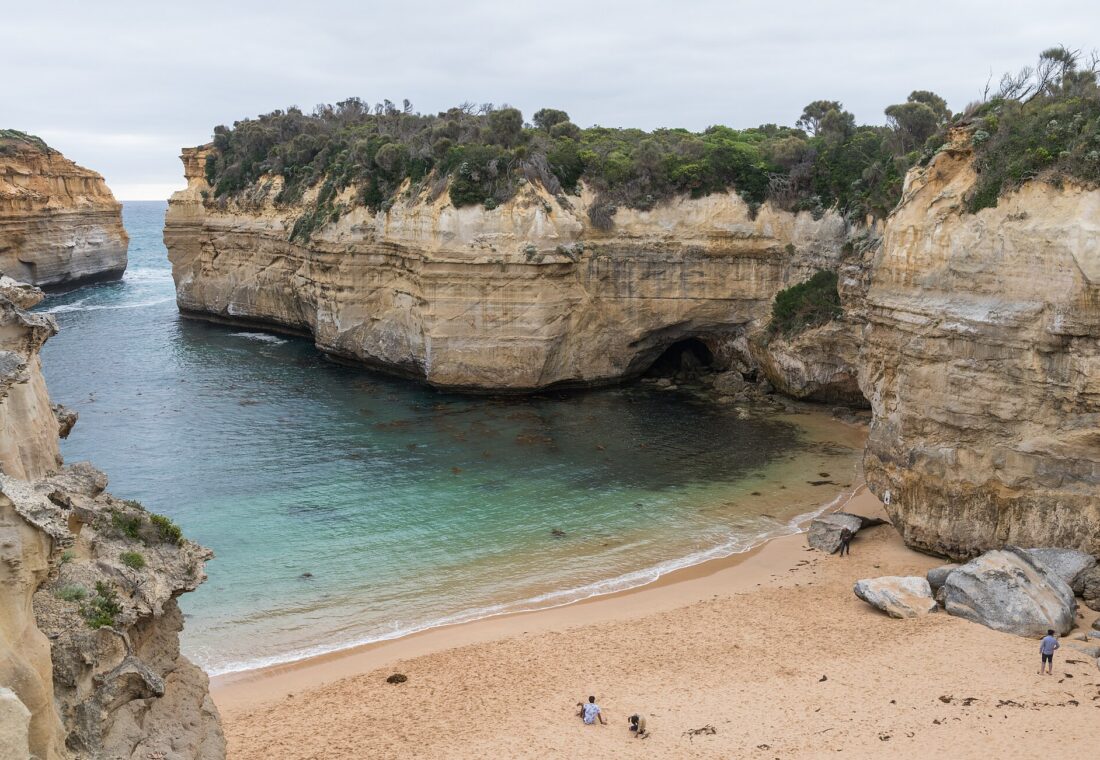 The sheltered sandy beach inside Loch Ard Gorge, enclosed by tall cream limestone cliffs topped with scrub, with calm green water and a few people on the sand