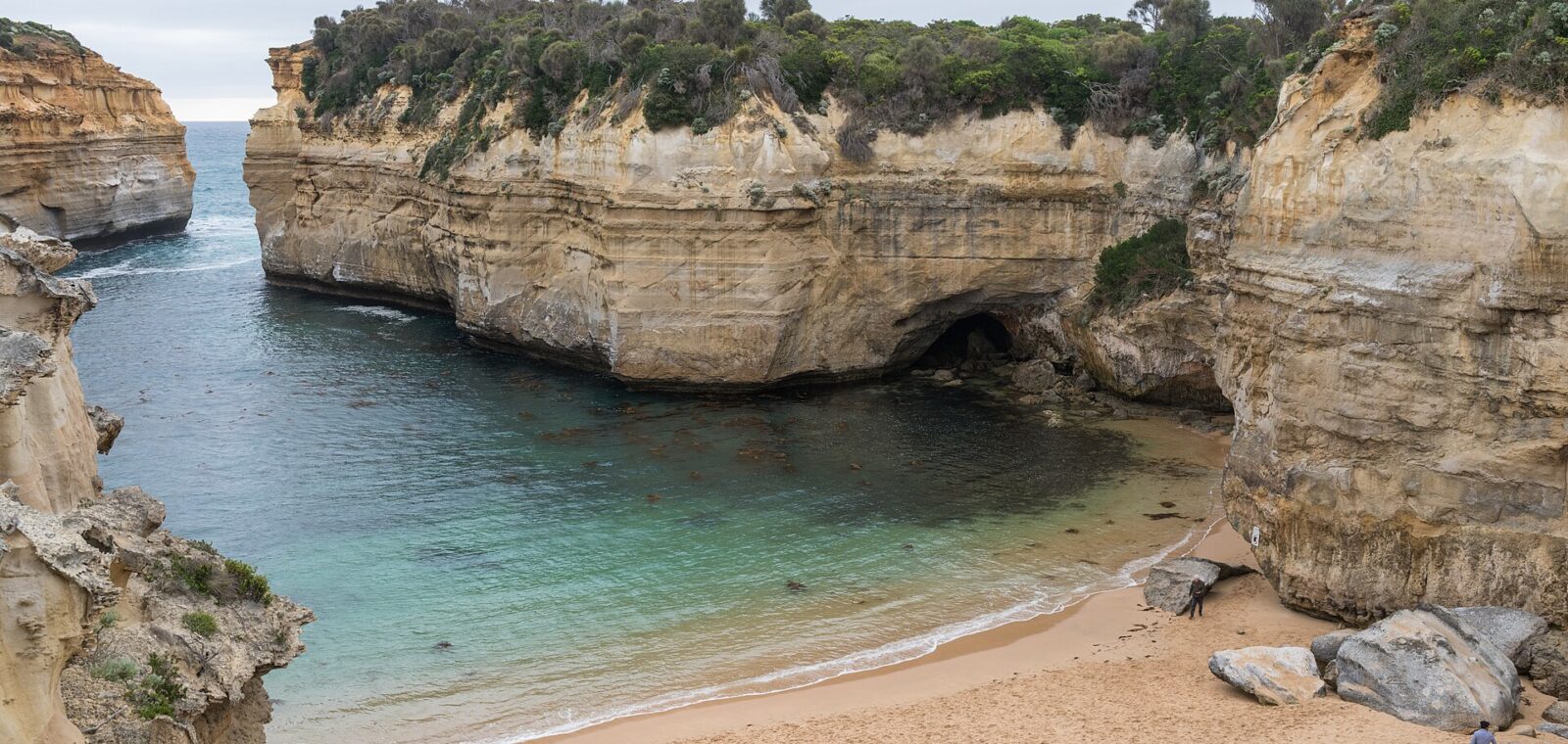 The sheltered sandy beach inside Loch Ard Gorge, enclosed by tall cream limestone cliffs topped with scrub, with calm green water and a few people on the sand