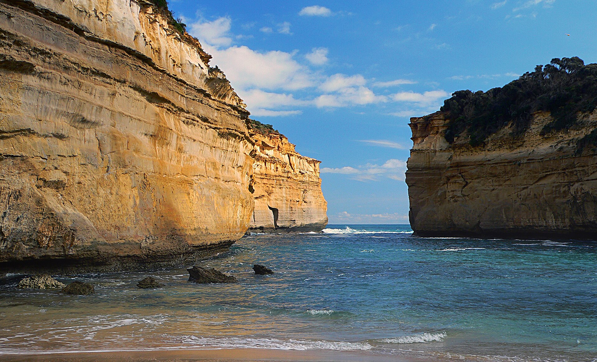 The narrow sea entrance to Loch Ard Gorge between two tall, layered limestone cliffs, with blue water and rocks in the foreground