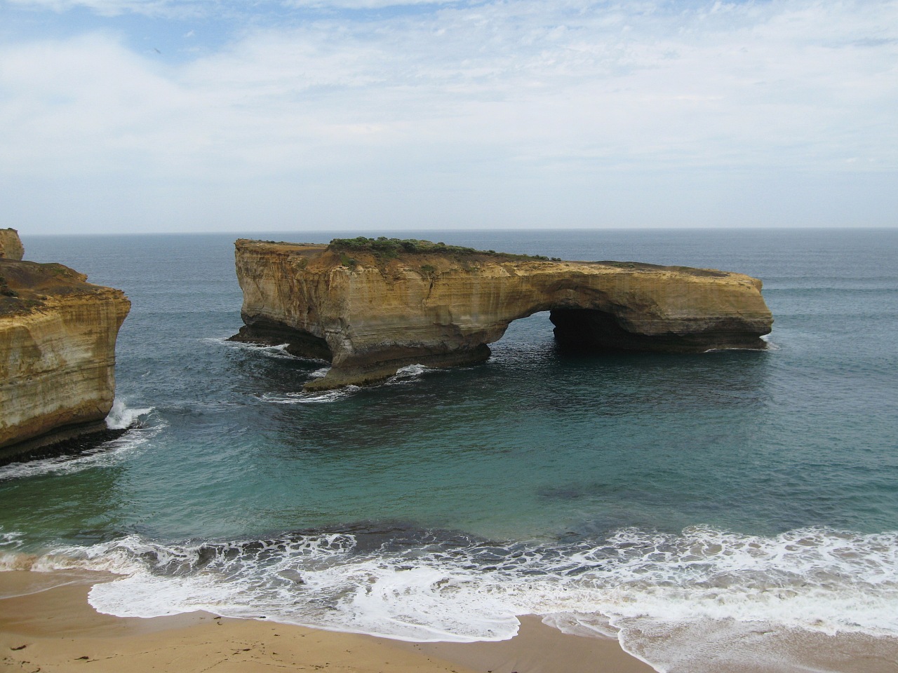 London Bridge, a flat-topped limestone stack with a single arch, standing offshore with waves breaking on the beach in the foreground