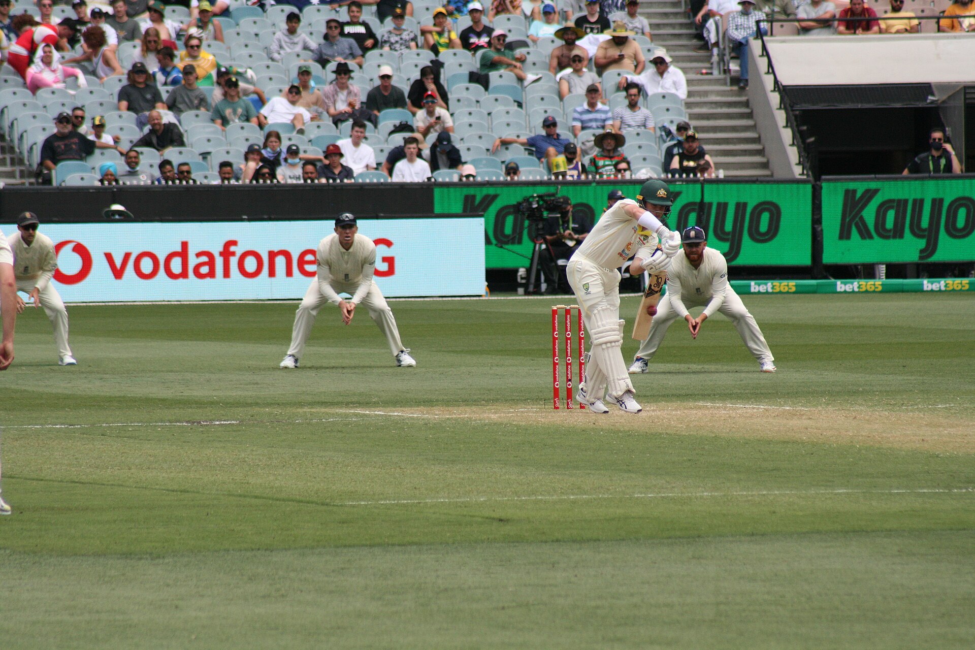 An Australian batter in whites plays a defensive shot as close fielders crouch around him at the MCG, with spectators in the stands behind