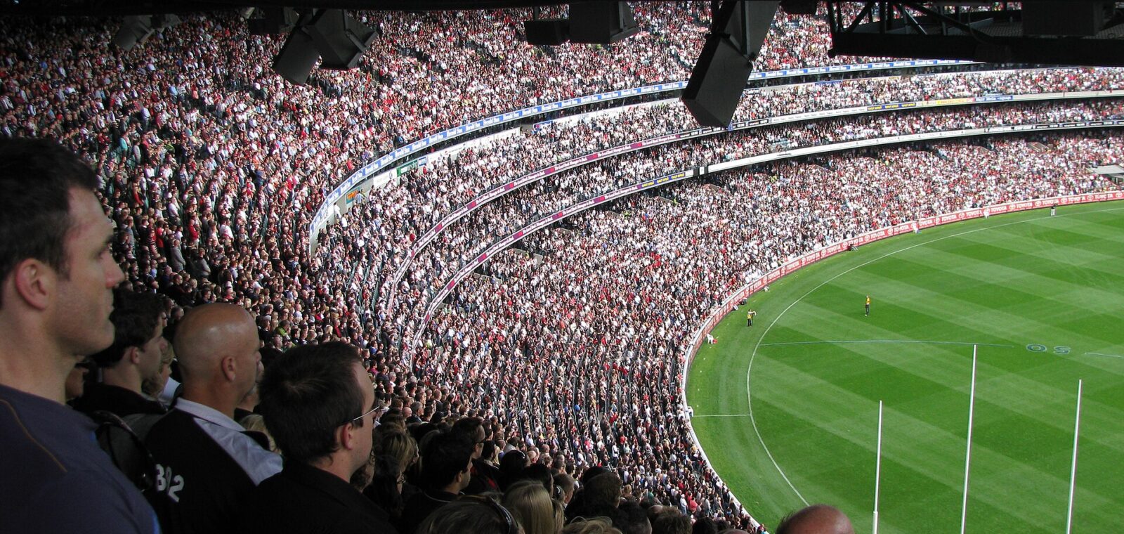 A packed MCG crowd watches an AFL match from the upper and lower decks, with the goalposts at the far end
