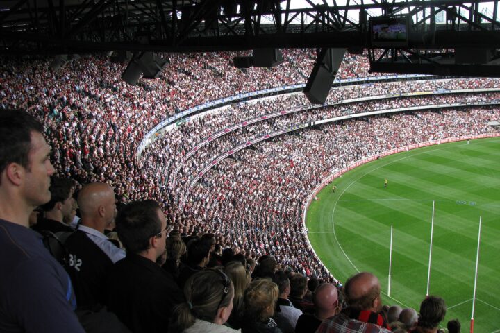 A packed MCG crowd watches an AFL match from the upper and lower decks, with the goalposts at the far end