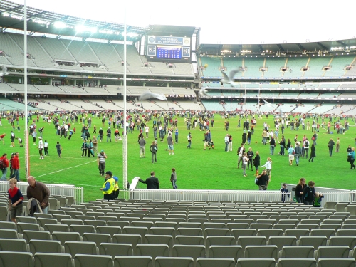 Fans of all ages kicking footballs on the MCG turf after a match, with the empty grandstands and goalposts behind
