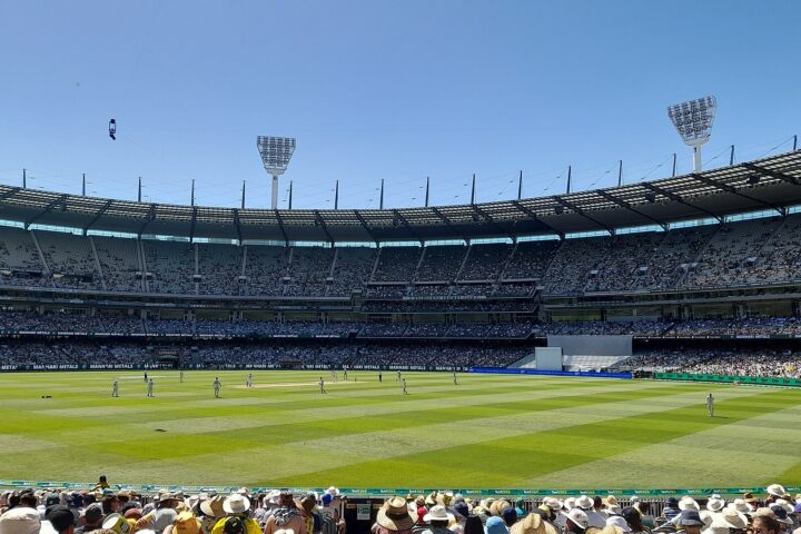 Spectators in hats in the MCG's Members' Reserve look out over a Test match on the green oval under a blue December sky