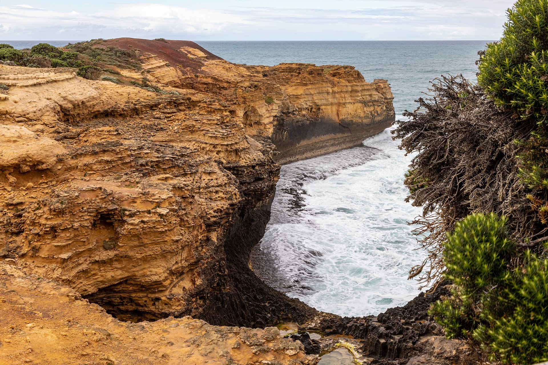 A sinkhole-like hollow in golden limestone at The Grotto, opening onto white surf and the ocean, with coastal shrubs on the right