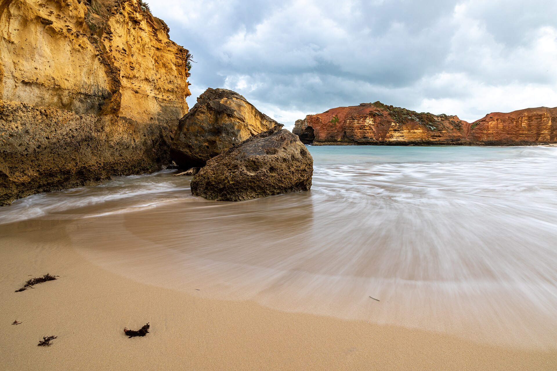 Waves washing over a sandy beach beside large limestone boulders and a cliff, with low ochre cliffs across the bay under a cloudy sky